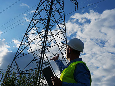 An energy engineer in white helmet, reflective vest, and holding a tablet, looks at an electric tower and wires. This energy engineer ensures the safety and efficiency of the power grid.